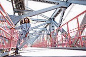 A dark-haired woman wearing sunglasses and a denim jumpsuit on a bridge