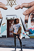 A dark-haired woman wearing a t-shirt, jeans and a jacket standing against a wall painted with street art
