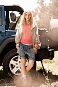 A blonde woman wearing a salmon-coloured jumper, a trenchcoat and shorts standing by a jeep