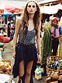 A brunette woman wearing a patterned top and a dark blue, dip-hem skirt at a market