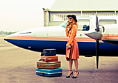 Brünette Frau in orangefarbenem Pullover und farblich passendem Rock, mit Koffern vor Flugzeug