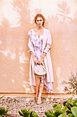 A blonde woman wearing a delicate pink summer dress and light coat standing against a wall