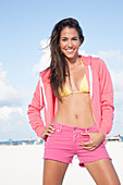 Woman wearing shorts and pink jacket over yellow bikini top standing at beach