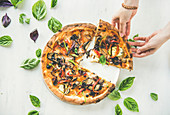 Female hands taking freshly baked Italian vegetarian pizza with vegetables and fresh basil over white marble table