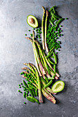 Variety of raw uncooked organic young green vegetables asparagus, peas, pod pea, avocado over grey texture background