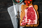 Raw fresh meat Club Steak, seasoning and meat fork and knife on dark wooden background