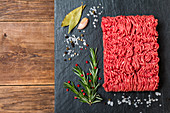 Minced meat on a slate stone black board with seasonings and fresh rosemary on wooden background, top view