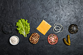 Ingredients for cooking black burger. Grilled chicken patty, buns, onion, tomatoes, lettuce, pickles, sauce, cheese over black background, top view