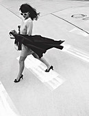 A dark-haired woman wearing a dress walking across the road holding a cape (black-and-white shot)