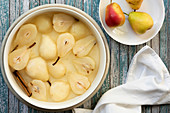 A bowl of poached pears and a cinnamon stick in syrup beside three uncooked red pears