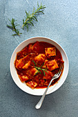 A bowl of ravioli in tomato and meat sauce with a fork on a blue textured background