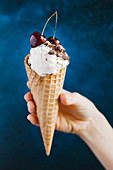 A hand holding vanilla ice cream, chocolate sprinkles and cherries in an ice cream cone