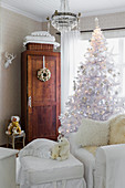 White Christmas tree and white loose-covered armchair with matching footstool in front of wooden cabinet