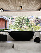 Dunkle, frei stehende Badewanne im Badezimmer mit Betondecke, Betonboden und Gartenblick