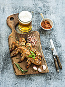 Fried chicken wings on rustic serving board, spicy tomato sauce, herbs and mug of light beer over black wooden backdrop
