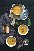 Red lentil soup with spices, herbs, bread in a rustic metal saucepan and bowls, over dark wood backdrop, top view, vertical