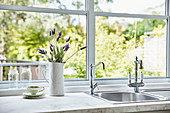 Jug of lavender flowers next to sink integrated in kitchen counter below window