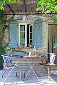 Table and metal chairs on wooden terrace below pergola outside window with blue shutters