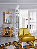 Gold-colored designer chair on carpet, mirror and bar trolley in matching colors in the background