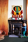 Kleinkind mit Roller vor antikem Kamin, Spielzeug-Dinosaurier und Wandbehang