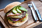 Burger with avocado slices, cheese, bacon, onions, lettuce, tomatoes on a wooden board with cutlery