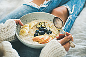 Woman in woolen sweater and jeans holding plate of vegan almond milk oatmeal porridge with berries, fruit and almonds