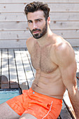 A young topless man sitting by a pool wearing shorts