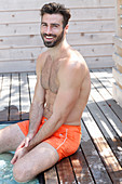 A young topless man sitting by a pool wearing shorts