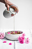 Almond, date, and cardamom granola with rosewater and rose petals, in bowl on linen with milk pouring