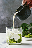 Cream pouring from jug to matcha green tea iced latte or cocktail in glass, with ice cubes, matcha powder on white marble table