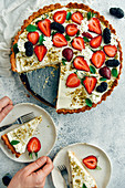 A woman serving a slice of strawberry cheese pie on a white plate, another slice and the rest of the pie accompany with lots of strawberries and mulberries