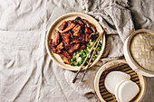 Chinese traditional dish Cantonese BBQ Pork Belly with spring onion served in ceramic plate with chopsticks, bamboo steamer and gua bao bus over linen cloth