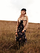 A blonde woman in the countryside wearing a hat, a dress and a brown jacket