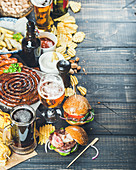 Variety of beers, grilled sausages, burgers, fried potato, corn, chips and sauces on dark wooden background