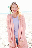 A blonde woman on a beach wearing a purple t-shirt and an apricot cardigan