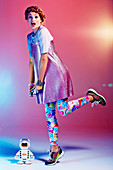 A girl with a plaited hairstyle wearing a glittery dress and colourful leggings playing with a toy robot