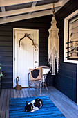 Stuhl vor weißer Tür, Makramee-Kronleuchter und Hund auf blauem Teppich auf Holzveranda