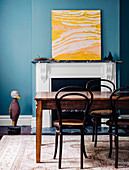 Wooden table with chairs in front of a fireplace, modern art in yellow tones on a mantelpiece