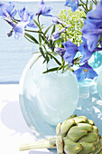 Blaue Ritterspornblütenin in Glasvase, Artischocke davor