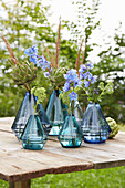 Blaue Glasvasen mit Artischocken und Rittersporn auf Holztisch im Garten