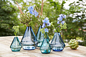 Blaue Glasvasen mit Artischocken und Rittersporn auf Holztisch im Garten