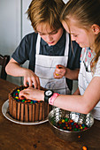 A boy and a girl decorating a chocolate cake wearing white aprons