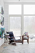 Vintage armchair and stacked books on side tables next to terrace doors and paintings on white wooden wall