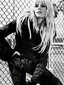 A blonde woman wearing a black jacket and trousers leaning against a fence (black-and-white shot)