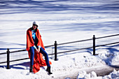 A brunette woman wearing a red cape, a blue shirt and blue trousers in a wintery park