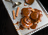 Cinnamon rolls with warm caramel topping and cinnamon sticks on a silver tray with tea-spoons