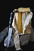 An entire mature cave aged cheddar cheese with its cloth rolled back and a slice removed on a dark rock with a vintage knife