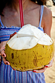 A fresh green coconut with a straw being held by a girl in a blue dress