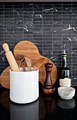 Kitchen utensils, wooden board and pepper mill against black-tiled splashback