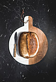 Kleiner Brotlaib mit Messer auf weiss-braunem Holzschneidebrett (Aufsicht)